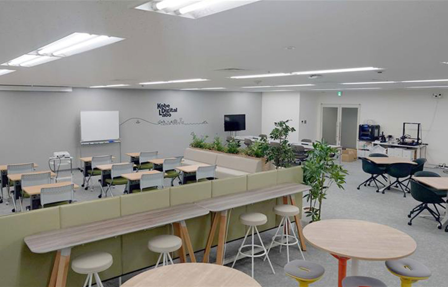ハイカウンターとスツール、丸テーブル、植栽の仕切りがあるオープンな共同スペースとその奥の教室エリア | Kobe Digital Labo。