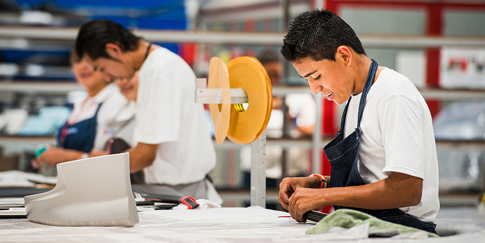 Homem de avental trabalhando com materiais em uma mesa de fabricação, com outras pessoas ao fundo.