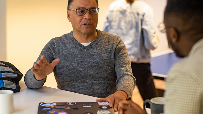 Pessoas reais, escritórios reais. Desenvolvedor masculino colaborando com um colega na sala de descanso ou cozinha do escritório. Colegas jogam pingue-pongue ao fundo. Um desenvolvedor personalizou seu laptop com adesivos.
