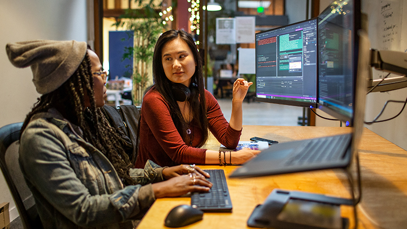 Duas desenvolvedoras trabalhando e colaborando em um escritório corporativo. Elas estão usando o Visual Studio com uma configuração de vários monitores.