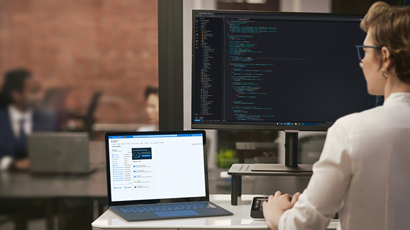 Uma mulher em frente a mesa olhando para um monitor com códigos e um notebook ao lado com pessoas ao fundo