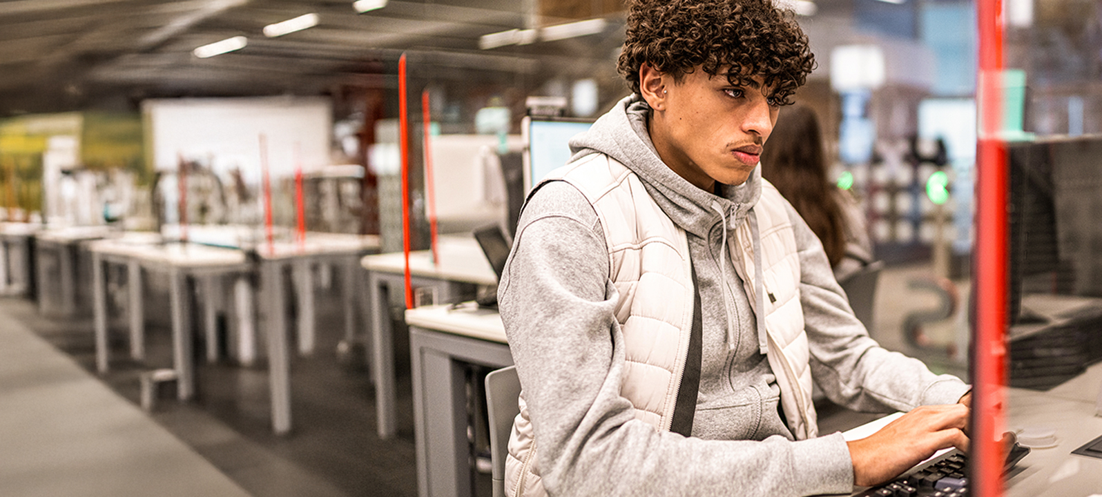 Um aluno está sentado em frente ao computador de uma biblioteca ou sala de aula, digitando no teclado. Fileiras de outras estações de computador são visíveis em segundo plano.