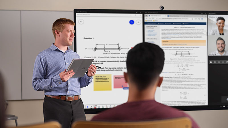 Uma pessoa fazendo uma apresentação com um tablet na mão, em pé na frente de uma tela grande exibindo diagramas, texto e uma interface de chamada de vídeo online.