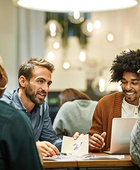 Um grupo de quatro pessoas senta-se ao redor de uma mesa, conversando. Duas delas estão olhando para um laptop, enquanto outra segura uma foto. Luzes suspensas e plantas são vistas ao fundo.