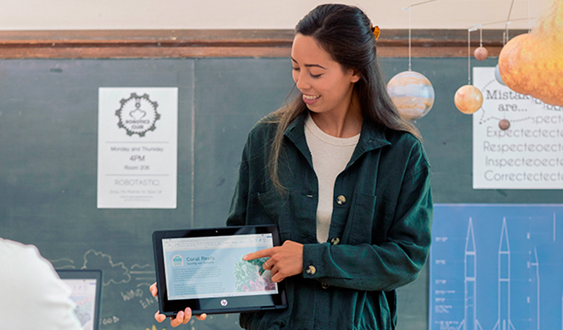 Uma mulher segura um tablet e aponta para a tela enquanto apresenta informações em uma sala de aula. Um quadro-negro com pôsteres e planetas pendurados está ao fundo. As costas de outra pessoa estão parcialmente visíveis.