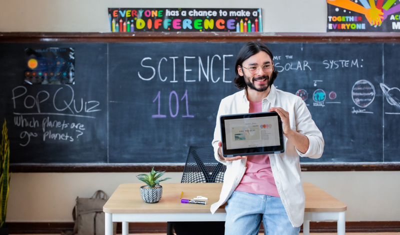 Um professor está em frente a um quadro negro da sala de aula com as palavras 'Ciências 101' escritas nele. Eles seguram um tablet exibindo um gráfico, provavelmente apresentando informações para a turma. Uma pergunta do questionário sobre planetas gasosos também está escrita no quadro. Um pequeno vaso de planta fica em primeiro plano na mesa do professor. Cartazes inspiradores estão pendurados acima do quadro negro.