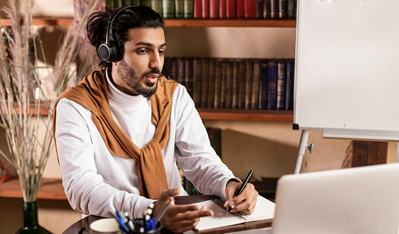 Um homem com fone de ouvido participa de uma videoconferência, fazendo anotações.