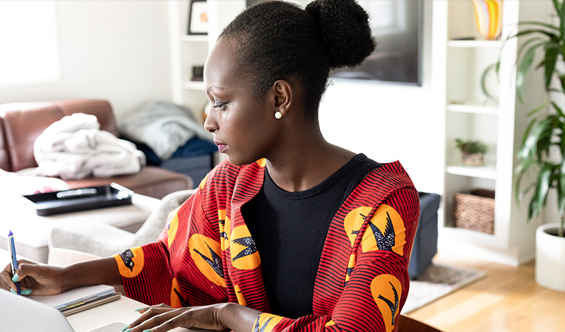 Uma mulher trabalha em casa, escrevendo em um caderno com um laptop por perto.