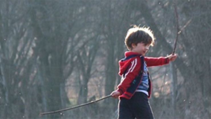 A kid in a forrest carrying a twig in each hand wearing a red jacket